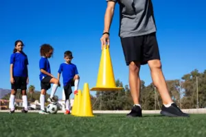 Foto ilustrativa de um auxiliar desportivo organizando material esportivo (bolas, cones, coletes) em uma quadra.