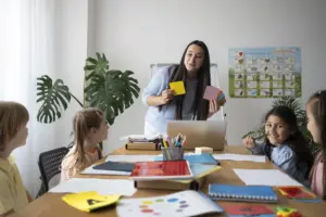 Foto ilustrativa de um auxiliar de sala interagindo com crianças em uma sala de aula.