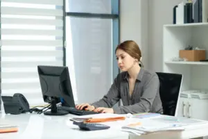 Assistente administrativo trabalhando em computador com documentos, ambiente de escritório público.