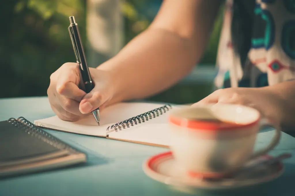 pessoa escrevendo em um caderno com xicara de café na frente