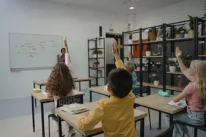 Alunos em sala de aula com professora ao fundo durante atividade. - Concurso SEE MG
