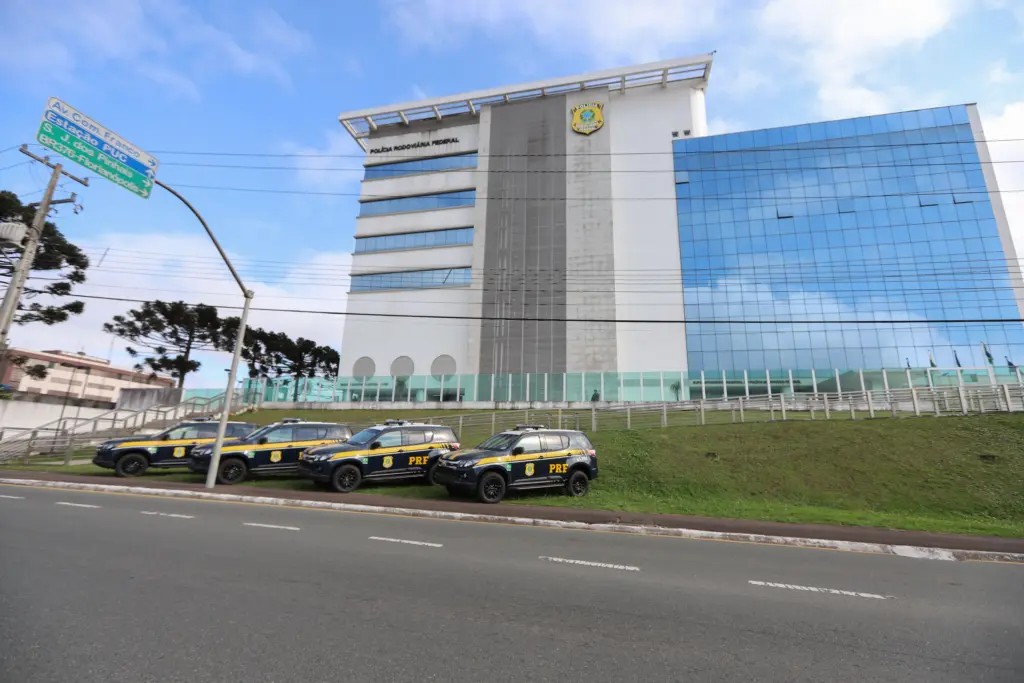 O que faz o Agente Administrativo da PRF? 1 Sede PRF com viaturas na frente e ao fundo céu azul com nuvens e araucárias. Agente administrativo da prf